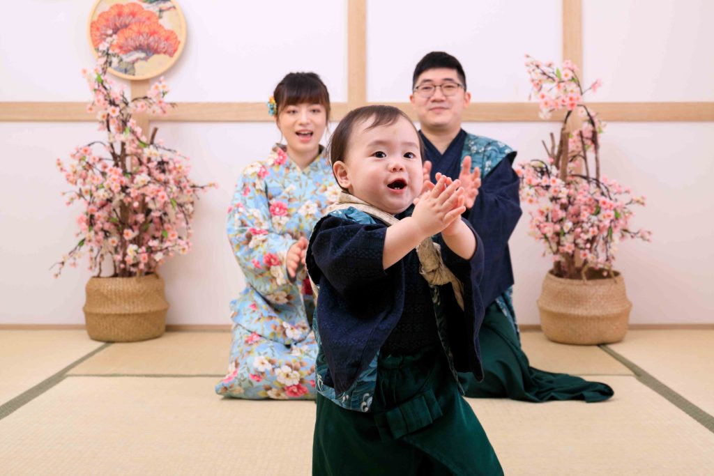 抓周儀式現場，滿周歲寶寶穿著和服開心拍手，父母在後方溫馨陪伴紀錄成長時刻。