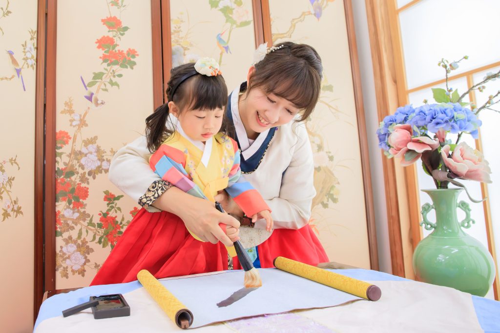 溫馨母女穿著韓服在傳統花鳥屏風前練習毛筆書法，媽媽手把手教小女孩揮毫，桌上擺有宣紙、墨盒與卷軸，旁邊放著插有藍色繡球花與粉色花朵的花瓶，呈現親子文化體驗與傳統藝術學習氛圍。