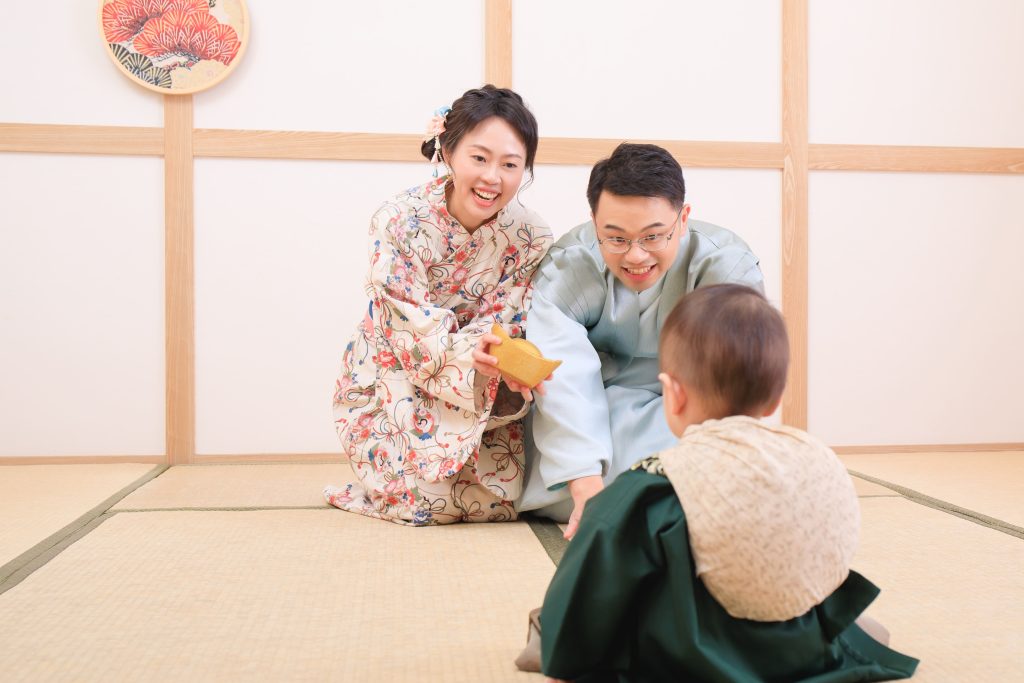 爸媽穿著和服，手拿著金元寶正鼓勵寶寶抓取，臉上表情興奮，象徵吉祥祝福的抓周活動場景。