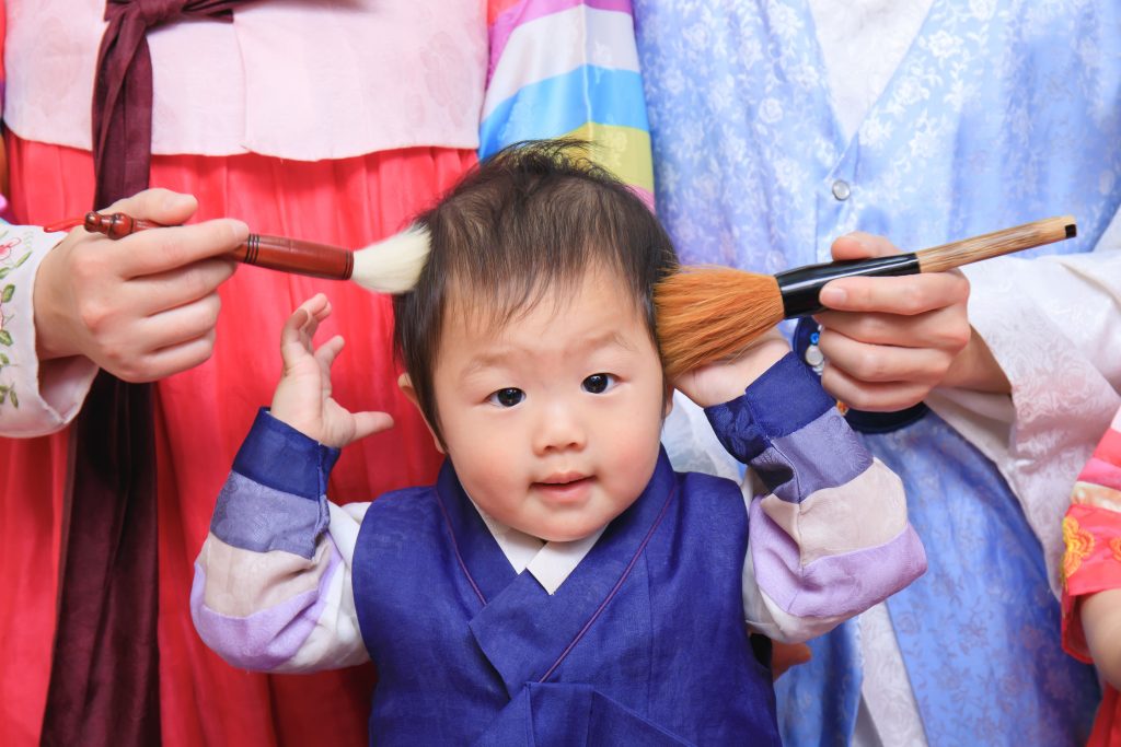 穿著韓服的寶寶參加抓周儀式，家人以毛筆輕掃臉頰，象徵智慧與祝福的溫馨畫面。