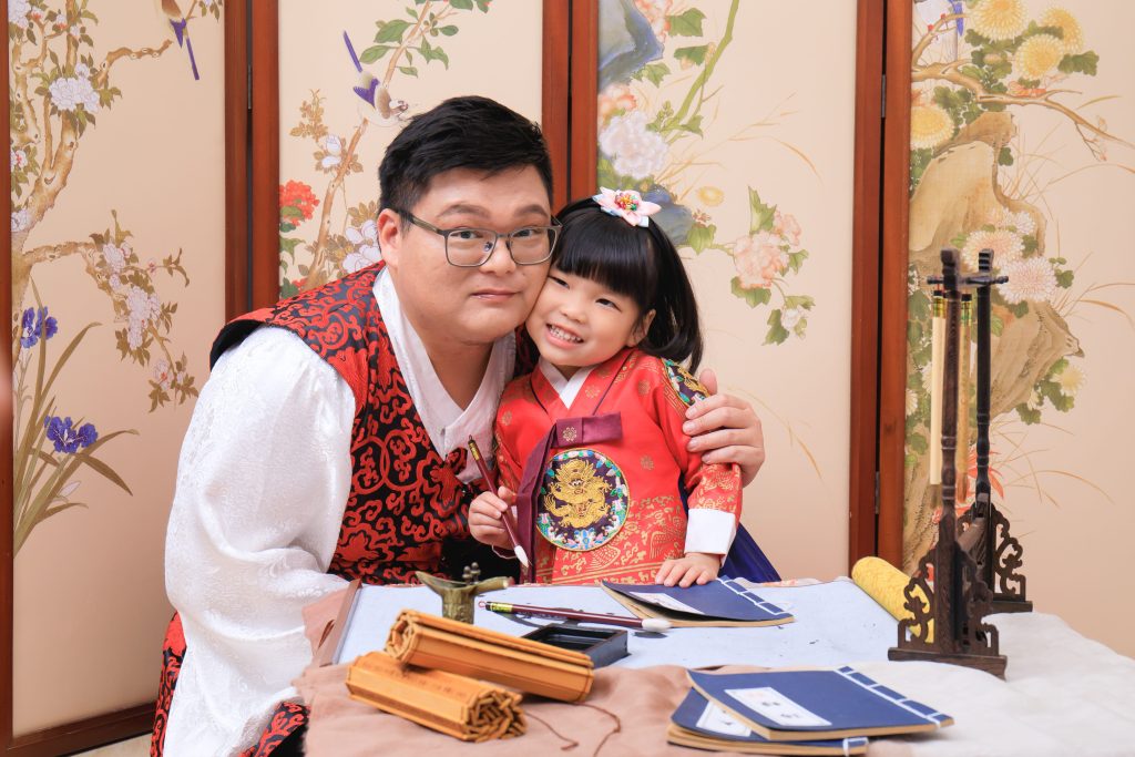 父女兩人穿著韓國傳統服飾，坐在書桌前，爸爸摟著女兒溫馨合影，展現深厚的親子情感。