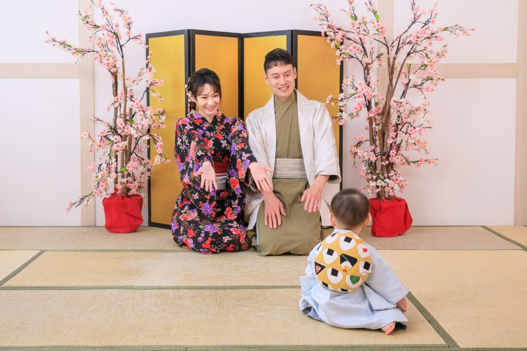 穿著日式和服的父母與寶寶參與傳統抓周儀式，寶寶背負象徵祝福的「一升餅」，背景為櫻花點綴和金色屏風，呈現溫馨的日式家庭氛圍。
