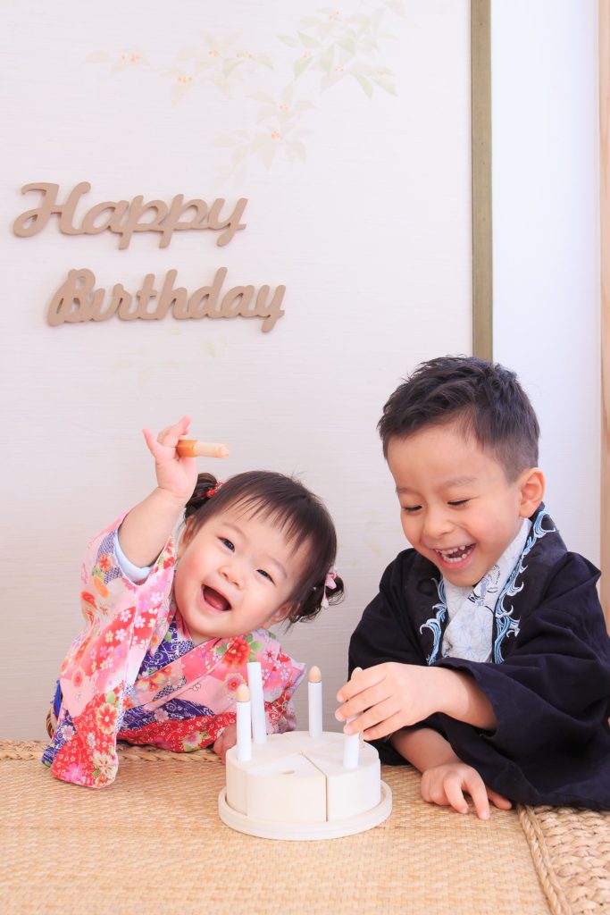 一歲寶寶抓周與生日紀念照，兄妹一起玩抓周道具，背景為 Happy Birthday 佈置，記錄寶寶的重要成長儀式