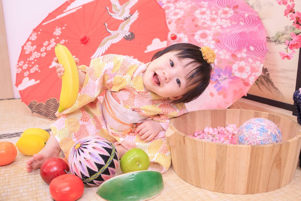 穿著日式傳統服飾的寶寶手握香蕉道具，坐在抓周場景中，背景有鮮豔的紅色和粉色和傘，象徵抓周活動的歡樂氣氛。