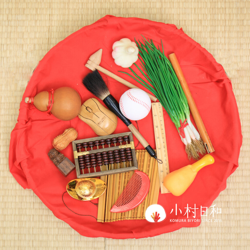 抓周物品全圖展示，包含毛筆、算盤、蔥、蒜、元寶、竹笛等象徵多種未來可能性的道具，擺放在紅色布上，充滿傳統儀式感與文化意義。