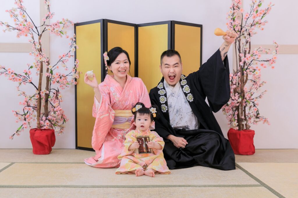日式家庭抓周儀式場景，父母身著日式和服，陪伴寶寶參與抓周活動，背景為櫻花點綴與金色屏風，捕捉溫馨愉快的家庭時光。